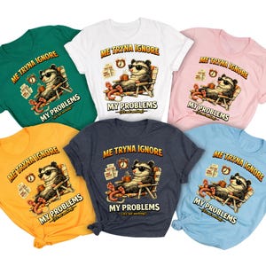 May include: A collection of six short-sleeve t-shirts in green, white, pink, yellow, dark gray, and light blue. Each shirt features a graphic of an opossum wearing sunglasses, lounging in a chair with a mug, and the text ME TRYNA IGNORE MY PROBLEMS.