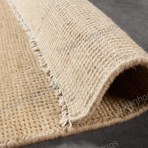May include: Close-up of a rolled-up beige rug with a textured, woven surface. The rug's edge is frayed, and the neutral color suggests a natural fiber. The rug is partially rolled, revealing its thickness and construction.