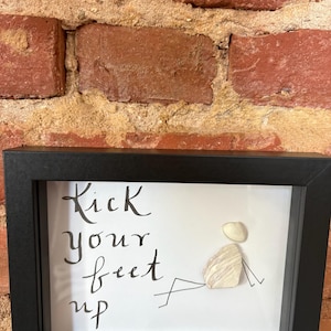 May include: A black framed wall art piece featuring the text "Kick your feet up" in black script. To the right, a figure is created using two white seashells for the head and body, with black ink lines depicting arms and legs in a reclining pose.