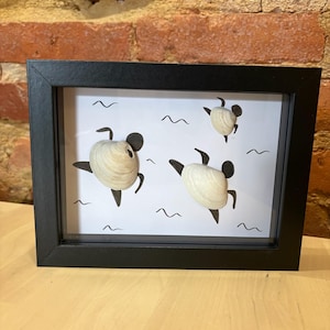 Può includere: Una cornice nera rettangolare a cassetta contenente uno sfondo bianco con tre figure create con conchiglie color bianco sporco e arti dipinti di nero. Le figure sono disposte in pose dinamiche, con piccole linee ondulate nere sparse intorno.