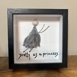 May include: A framed artwork featuring a shell angel with a halo, wings, and a shell dress. The artwork is set against a white background, with the phrase "pretty as a princess" written in black script. The frame is black.