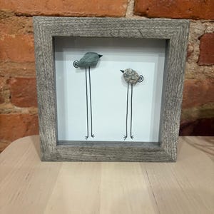 Könnte beinhalten: Ein gerahmtes Kunstwerk mit zwei skurrilen Vögeln. Die Vögel sind aus Steinen gefertigt, mit dünnen schwarzen Beinen und einem minimalistischen Design. Der Rahmen ist verwittert grau, was dem Stück eine rustikale Note verleiht. Der Hintergrund ist weiß.