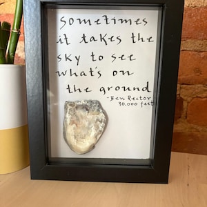 May include: Black framed art print featuring the quote "Sometimes it takes the sky to see what's on the ground" by Ben Rector. The print includes a natural, off-white, textured object. The frame is rectangular.