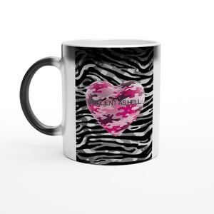 Op de afbeelding: Witte keramische mok met een zwart handvat. De mok heeft een roze camouflage hart met de tekst "RESILIENT AS HELL" tegen een zwart-witte zebraprint achtergrond.
