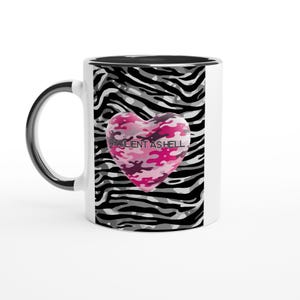 Op de afbeelding: Witte keramische mok met een zwart handvat en rand. De mok heeft een zebraprint ontwerp met een roze camouflage hart met de tekst "RESILIENT AS HELL".