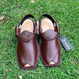 May include: A pair of brown leather Peshawari sandals with a strap and buckle closure. The sandals have a light-colored insole and are displayed on a bed of green grass. A tag with the text "SoleMehal" is attached.