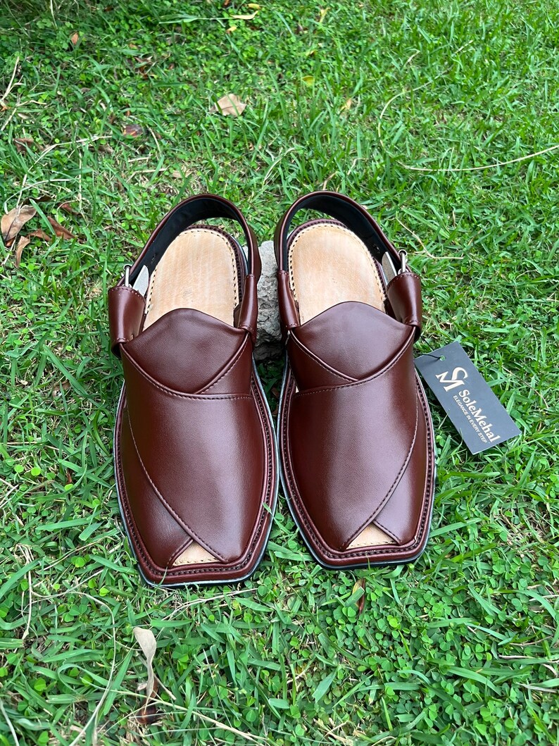 May include: A pair of brown leather Peshawari sandals with a strap and buckle closure. The sandals have a light-colored insole and are displayed on a bed of green grass. A tag with the text "SoleMehal" is attached.