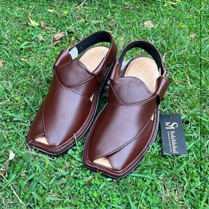 May include: A pair of brown leather sandals with a unique design, featuring a strap around the ankle. The sandals are placed on a bed of green grass. A tag with the text "SoleMehal" is attached to one sandal.
