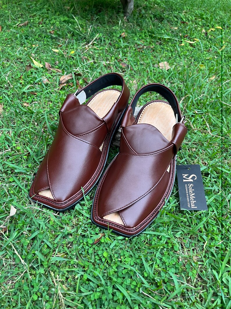 May include: A pair of brown leather sandals with a unique design, featuring a strap around the ankle. The sandals are placed on a bed of green grass. A tag with the text "SoleMehal" is attached to one sandal.