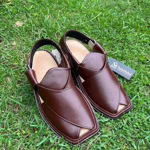 May include: A pair of brown leather Peshawari sandals with a square toe and a strap around the heel. The sandals are set on a bed of green grass. The sandals have a light brown interior and a black sole.