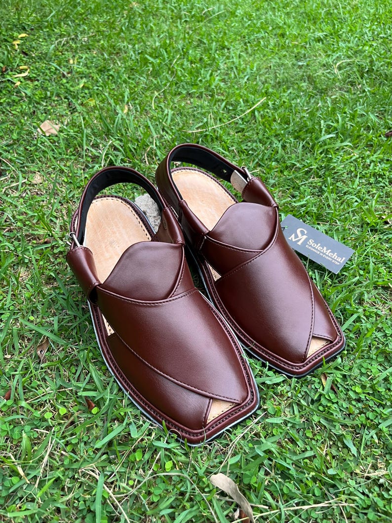 May include: A pair of brown leather Peshawari sandals with a square toe and a strap around the heel. The sandals are set on a bed of green grass. The sandals have a light brown interior and a black sole.