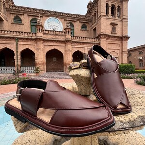 May include: A pair of brown leather sandals with a buckle closure, displayed on a stone surface. The sandals have a classic design with open sides and a closed toe. The background features a brick building.