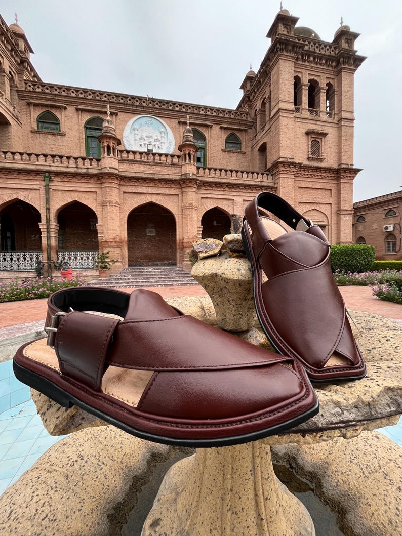 May include: A pair of brown leather sandals with a buckle closure, displayed on a stone surface. The sandals have a classic design with open sides and a closed toe. The background features a brick building.