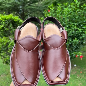 May include: A pair of brown leather Peshawari sandals with a square toe design. The sandals have a strap around the ankle and a light-colored insole. The edges are trimmed with a white and black border. The background is a blurred green.