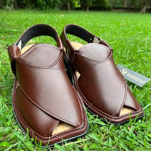 May include: A pair of brown leather sandals with a cross-strap design, set on a bed of green grass. The sandals have a black sole and a strap around the ankle. A small tag with text is visible.