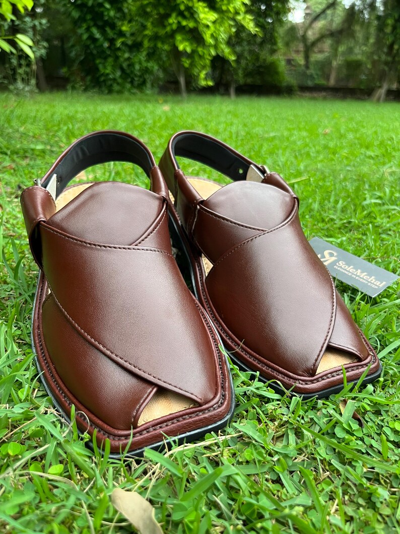 May include: A pair of brown leather sandals with a cross-strap design, set on a bed of green grass. The sandals have a black sole and a strap around the ankle. A small tag with text is visible.