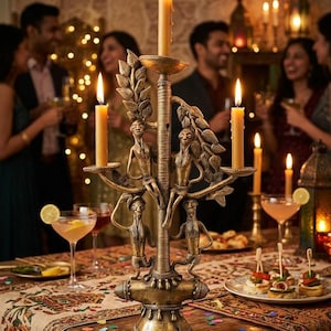 May include: An ornate brass candelabra with four lit candles. The candelabra has a central column with human-like figures and leaf detailing. The scene is set on a table with a patterned tablecloth, drinks, and appetizers, suggesting a social gathering.