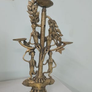 May include: A brass sculpture depicting a tree with four figures and leaf detailing. The sculpture features a central column with a dish at the top and two side platforms. The figures are arranged around the central column.