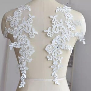 May include: Two symmetrical white lace appliques featuring intricate floral and vine embroidery patterns. The delicate needlework displays detailed petals, leaves, and swirling stems, designed to be placed on fabric for decorative embellishment.