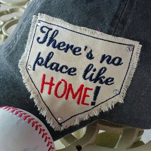 May include: A black baseball cap with a white patch that says "There's no place like HOME!" in blue and red embroidery. The patch has a diamond shape and is decorated with rhinestones.