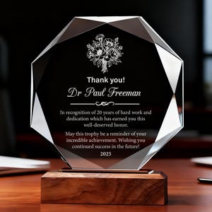 Può includere: Un trofeo di cristallo trasparente e sfaccettato con base in legno. Il trofeo presenta il testo "Thank you! Dr Paul Freeman" e un motivo floreale. Il testo continua con un messaggio di riconoscimento e l'anno 2025.