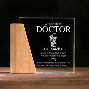 Könnte beinhalten: Transparente Glasplakette mit schwarzem Hintergrund und Holzsockel. Auf der Plakette steht "A TRULY GREAT DOCTOR" mit Stethoskop und Blumendesign. Enthält einen Namen und eine Nachricht. Der Sockel ist hellbraun.