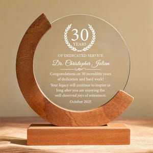 Puede incluir: Un premio de vidrio transparente con una base de madera y un acento de madera curvado. El premio presenta el texto "30 años de servicio dedicado" y un mensaje personalizado. El premio es un regalo de jubilación.