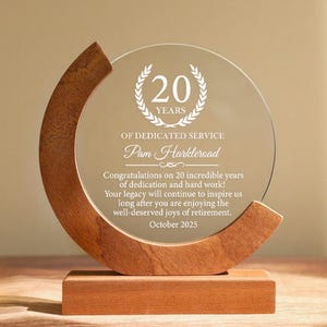 May include: A clear glass award with a wooden base and a curved wooden accent. The award commemorates 20 years of service and includes the name "Pam Harkleroad" and the date "October 2025".
