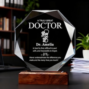 Könnte beinhalten: Eine Kristallauszeichnung mit schwarzem Hintergrund und Holzsockel. Der Text lautet "A Truly Great Doctor" mit einem Stethoskop und floralem Design. Darunter steht "Dr. Amelia" und ein Zitat. Ein nachdenkliches Geschenk für einen Arzt.
