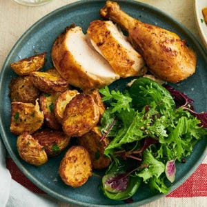 May include: A plate of roasted chicken with sliced breast, a drumstick, and roasted potatoes. A fresh green salad with red accents is also on the plate. A glass of yellow liquid and a plate of potatoes are in the background.