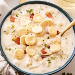 Puede incluir: Un cuenco de sopa cremosa blanca, adornada con galletas redondas y trozos de tocino. Una cuchara dorada descansa en la sopa, y se ve una copa de vino blanco. La sopa está sobre una superficie blanca.