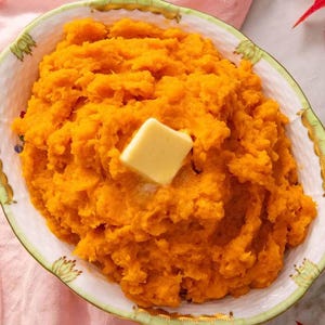 Könnte beinhalten: Eine Schüssel mit leuchtend orangefarbenem Süßkartoffelpüree, garniert mit einem Butterstück. Die Schüssel hat einen dekorativen Rand mit einem grün-weißen Design. Ein rosa Tuch und Herbstblätter sind im Hintergrund.