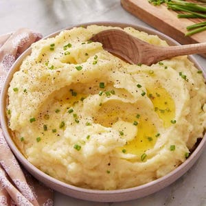 Puede incluir: Un cuenco de puré de patatas cremoso, adornado con cebollino y rociado con aceite. Una cuchara de madera descansa en el centro. El plato está sobre una mesa con un cuenco pequeño de pimienta, cebollino y un jarrón con flores.