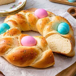May include: A braided bread wreath decorated with pastel-colored eggs and sprinkles. A slice of the bread is cut, revealing the soft interior. Additional eggs in a wooden holder and a plate of bread slices are in the background.