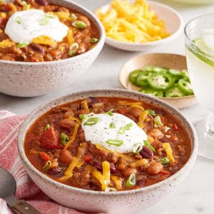 May include: A close-up shot of a hearty chili dish in a speckled bowl, topped with sour cream, shredded cheese, and green onions. Additional bowls contain cheese, jalapeños, and tortilla chips. A refreshing drink with lime slices is also visible.