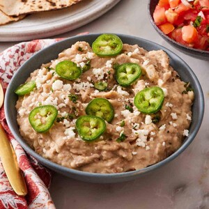 May include: A bowl of refried beans topped with sliced jalapeños and crumbled cheese. Tortillas, pico de gallo, and a bowl of cheese are also visible, suggesting a Mexican-inspired meal.