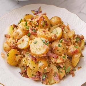 Puede incluir: Ensalada de patatas con bacon y hierbas en un plato blanco con borde dorado y diseños de mariposas. Un cuenco de ensalada de patatas en el fondo.