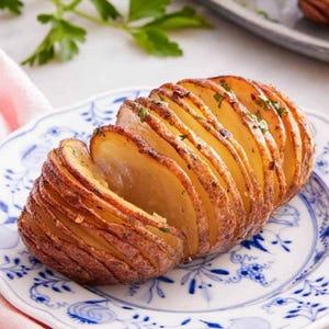 May include: A hasselback potato, sliced and baked, on a blue and white patterned plate. Fresh herbs are visible. Other baked potatoes are on a gray platter in the background. The image shows a close-up.