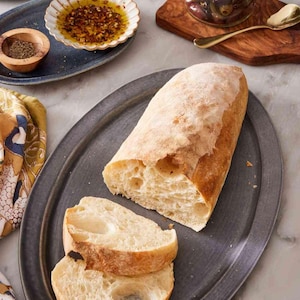 Puede incluir: Un pan crujiente parcialmente rebanado sobre una fuente ovalada gris oscuro. Se ven dos rebanadas. Los platos incluyen aceitunas, aceite de oliva con especias y sal. También hay una cuchara dorada.