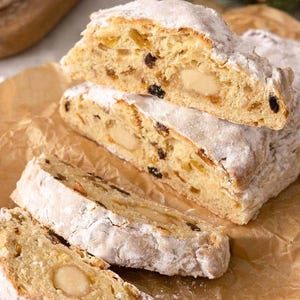 Puede incluir: Primer plano de rebanadas de stollen espolvoreadas con azúcar glas, que revelan almendras, pasas y frutas confitadas. El pastel está sobre papel de pergamino marrón.