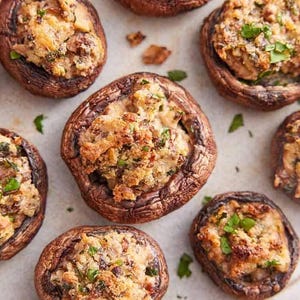 Può includere: Vista dall'alto di un piatto con diversi funghi ripieni. I funghi sono marroni con un ripieno di colore chiaro e guarniti con erbe verdi. Il piatto sembra un antipasto saporito.