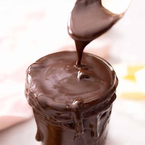 May include: A close-up shot of a jar being drizzled with rich, dark chocolate sauce. The glossy sauce cascades from a spoon, coating the top and sides of the jar. The image highlights the texture and color of the chocolate.