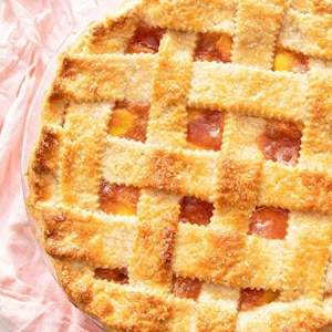 May include: A homemade peach pie with a lattice pastry top. The golden-brown crust is dusted with sugar, and the filling is a bright orange hue. The pie is in a clear glass dish, and rests on a pink cloth.
