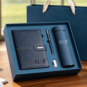 May include: A dark blue gift set containing a notebook, a pen, and a cylindrical bottle, all featuring the monogram G|F. The items are presented in a matching dark blue box and a gift bag with light-coloured ribbon handles, set against a blurred background.