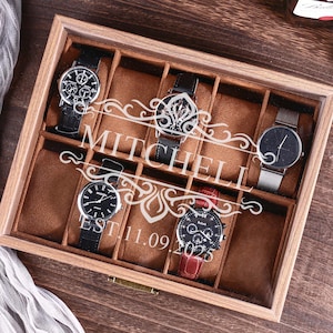 May include: A wooden watch display case with six compartments, each holding a wristwatch. The case is personalized with the name "MITCHELL" and the date "EST 11.09.2025". The watches have black, silver, and red bands.