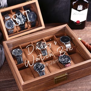 May include: A wooden watch box with a glass top, holding several wristwatches. The box is engraved with "Daddy's Watch Box" and "Love, James". The watches have various band and face designs.