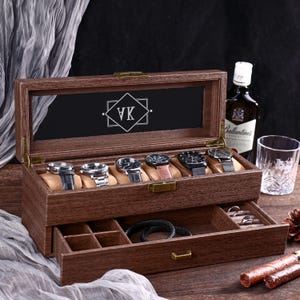 Puede incluir: Una caja de reloj de madera oscura con tapa de cristal, que muestra seis relojes sobre cojines color canela. La caja tiene un cajón para almacenamiento adicional. La parte superior de la caja tiene las iniciales "VK" en forma de diamante. Una botella de whisky y un vaso están en el fondo.