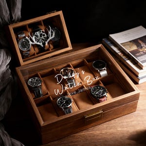 Pode incluir: Uma caixa de relógios de madeira com tampa de vidro, exibindo vários relógios de pulso. A caixa é gravada com "Daddy's Watch Box, love, James". Os relógios têm vários designs de pulseira e mostrador. A caixa está aberta, revelando vários compartimentos.