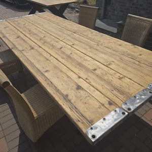 Könnte beinhalten: Ein rechteckiger Esstisch aus hellbraunen Holzplanken mit sichtbarer Maserung und Astlöchern. Der Tisch verfügt an den Enden über silberne Metall-Eckbeschläge im Industriestil mit sichtbaren Schraublöchern. Ein Logo zeigt OMH WOODS & MORE.