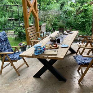 Könnte beinhalten: Ein rustikales Ess-Set für den Außenbereich mit einem langen, hellen Holztisch mit schwarzen Metallbeinen. Zwei Holzstühle mit blau gemusterten Kissen befinden sich auf beiden Seiten. Der Tisch ist mit Tellern, Gläsern und Essen gedeckt, unter einer Holzpergola.
